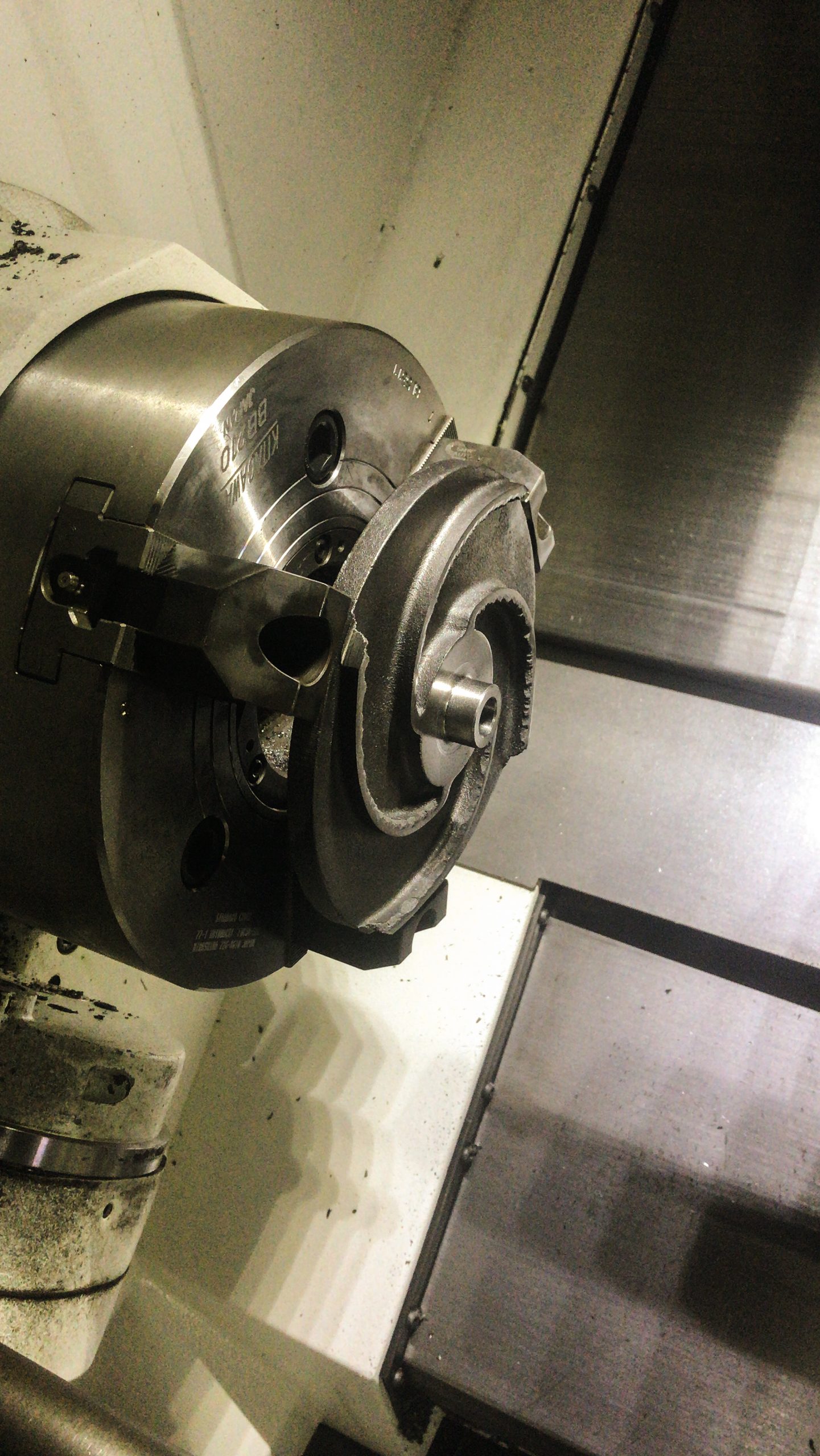 Versleten metalen pompwaaier ingespannen in een draaibank voor bewerking, met machineonderdelen en metaalspanen in een werkplaatsomgeving.