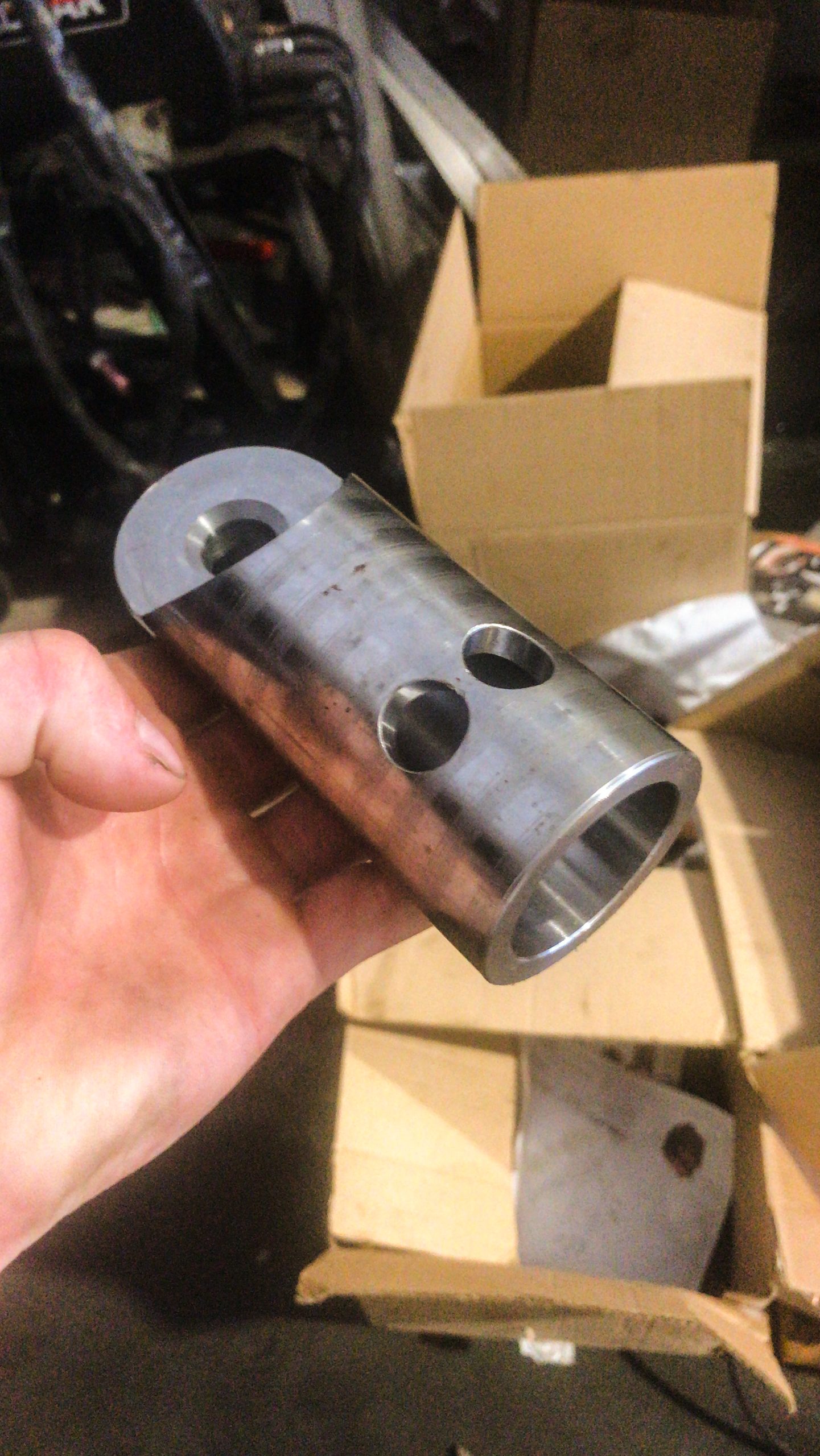 Hand houdt een bewerkte metalen huls met meerdere boorgaten, met kartonnen dozen en werkplaatsmateriaal op de achtergrond.