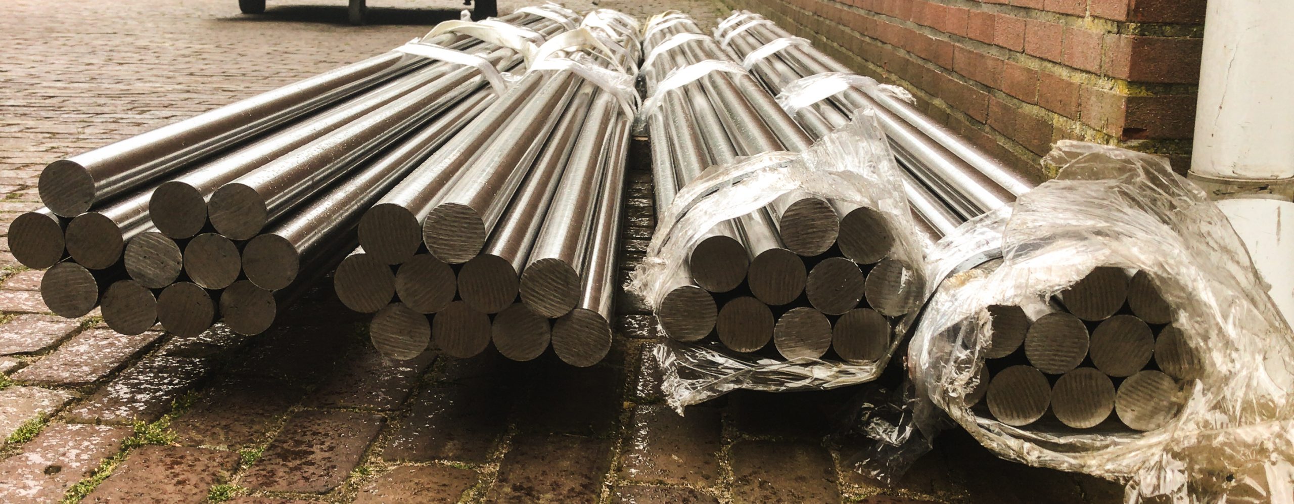 Strak gebundelde roestvrijstalen staven, verpakt in plastic en gestapeld op een natte bestrating naast een bakstenen muur.
