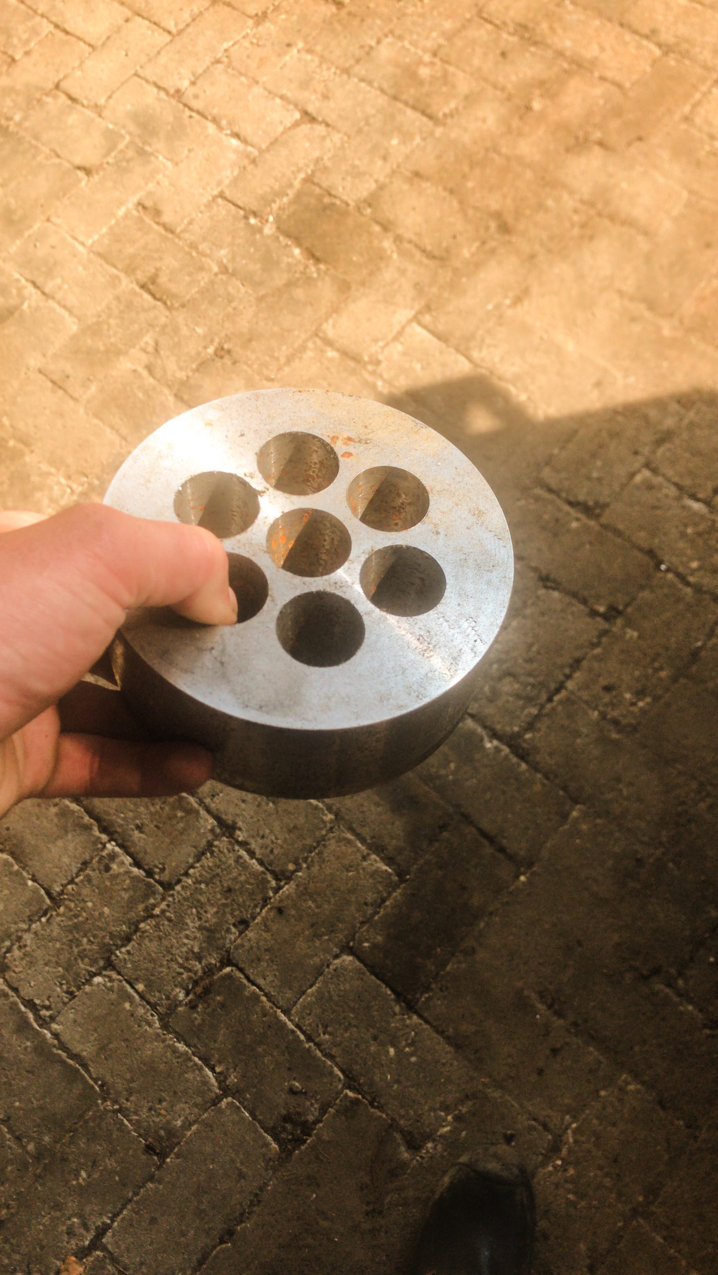 Hand houdt een ronde metalen schijf met zeven geboorde gaten, tegen een achtergrond van een bestrate werkplaatsvloer.