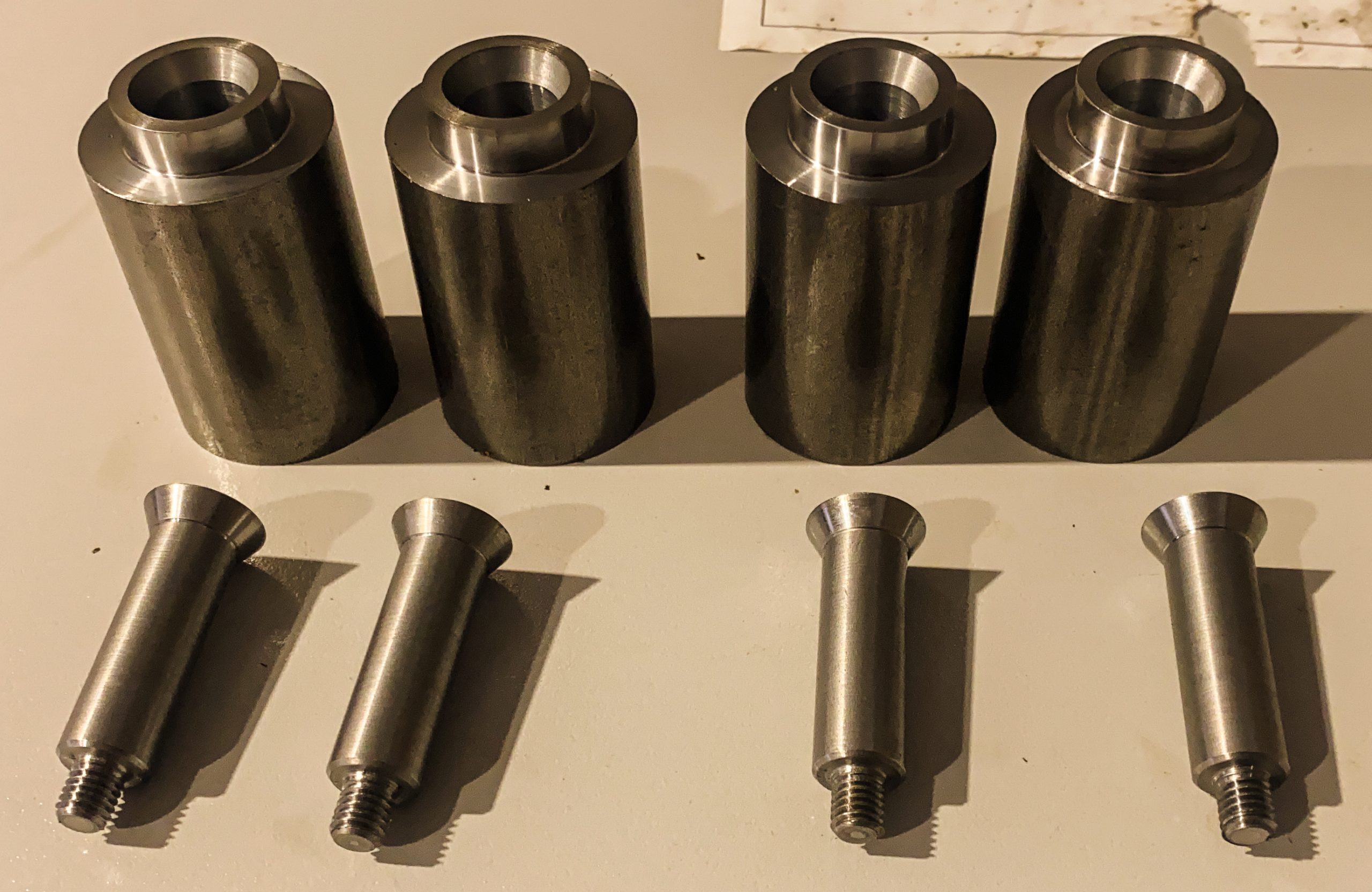 Vier bewerkte metalen cilinders en vier boutvormige metalen onderdelen met schroefdraad, netjes gerangschikt op een werkblad.
