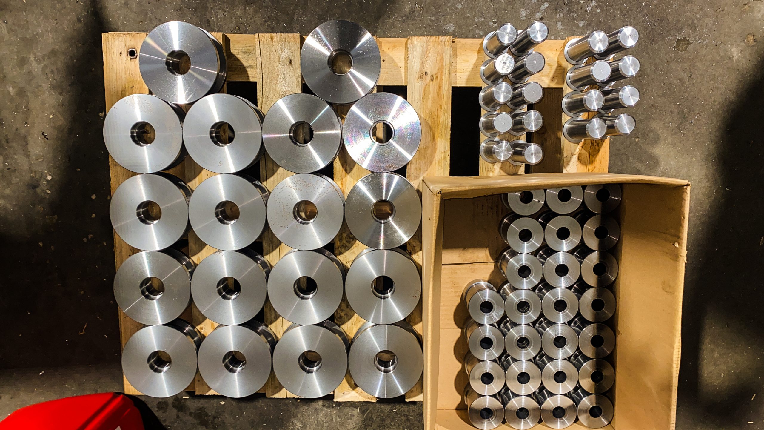 Stapel bewerkte metalen schijven en cilinders geordend op een houten pallet en in een kartonnen doos in een werkplaats.